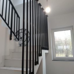 Stal Zachodniopomorskie - Białe schody z czarną, minimalistyczną balustradą, na ścianie dekoracyjny zegar, widok na zewnątrz przez okno.