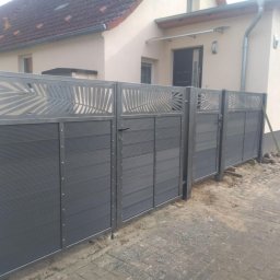 Stal Zachodniopomorskie - Nowoczesne ogrodzenie panelowe w kolorze grafitowym z dekoracyjnymi wycięciami w kształcie liści palmowych, zamontowane przed domem z beżową elewacją i brązowym dachem.