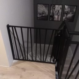 Stal Zachodniopomorskie - Nowoczesna, czarna barierka schodowa z ukośnymi prętami, zamontowana wewnątrz domu na jasnej podłodze, z widocznymi schodami i trzema czarno-białymi obrazami na szarej ścianie w tle.