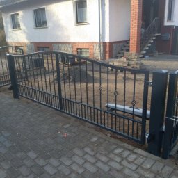 MJ - Automatyczna brama wjazdowa z czarnego metalu, z dekoracyjnymi elementami, zamontowana przed domem w trakcie budowy, widoczny mechanizm otwierający.