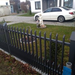 Stal Zachodniopomorskie - Nowoczesne ogrodzenie z metalowych sztachet o skośnych zakończeniach, widok na trawnik, rzeźbę ogrodową i biały samochód zaparkowany przed domem.