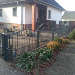 Metalowe ogrodzenie posesji z bramą wjazdową, widoczne stosy kostki brukowej przygotowane do dalszych prac wykończeniowych.