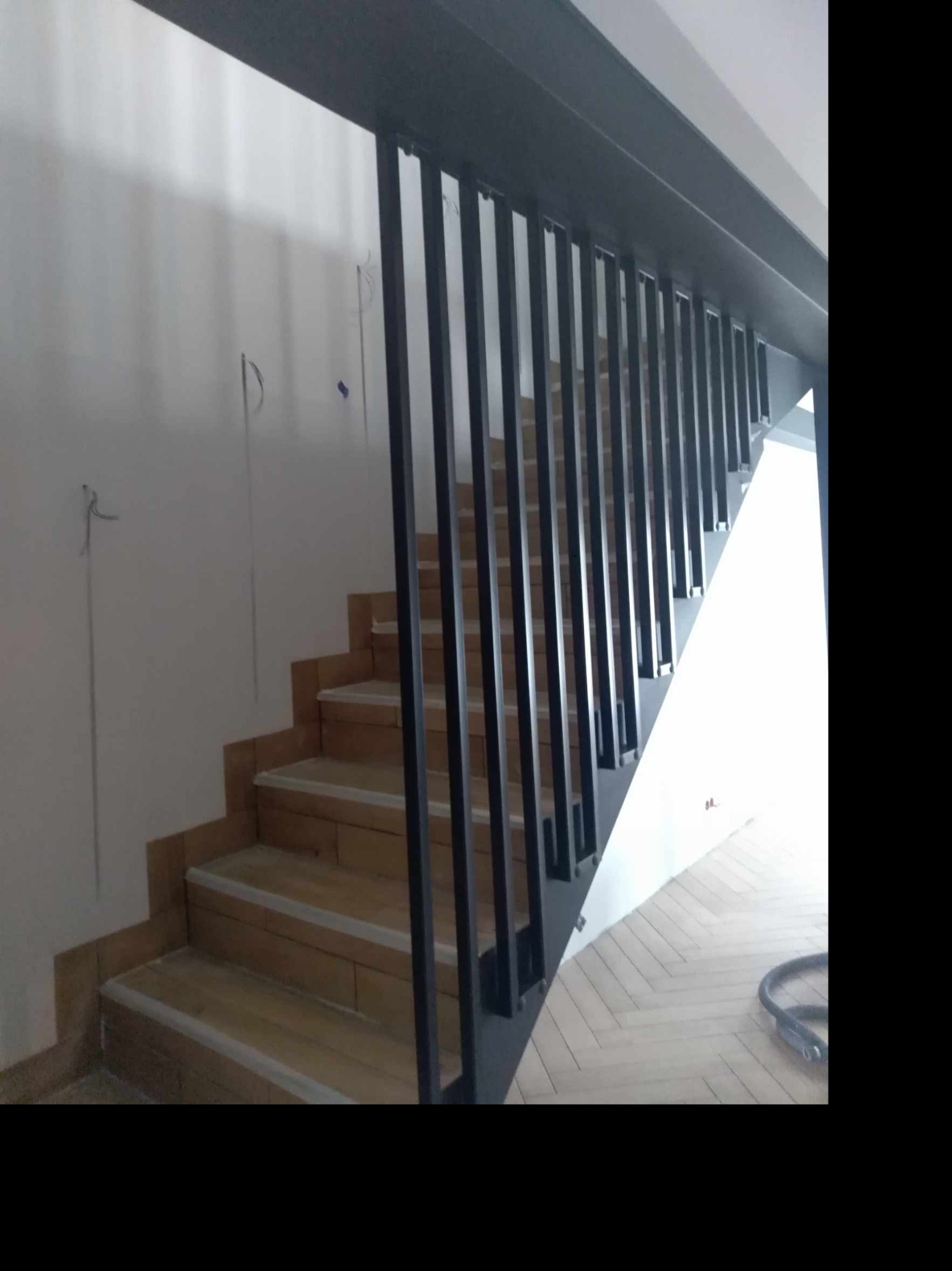 Metalowa balustrada z pionowymi szczeblami w kolorze grafitowym, zamontowana przy schodach wyłożonych terakotą z białymi paskami, widok z dołu na jasne, parkietowe podłoże.