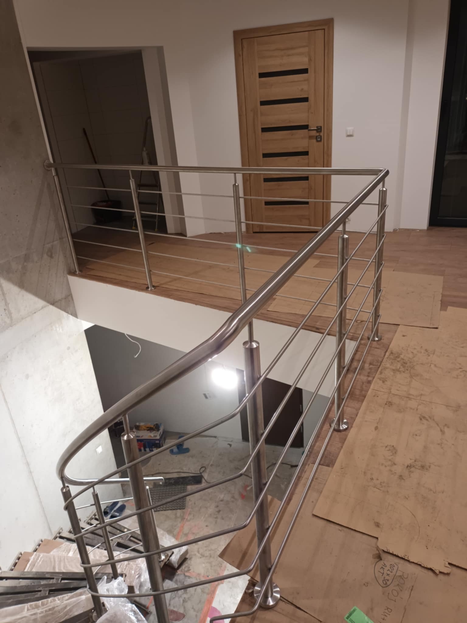 Wnętrze w trakcie wykańczania: widok z góry na schody z balustradą ze stali nierdzewnej, zabezpieczone podłogi kartonem, drzwi drewniane w tle.