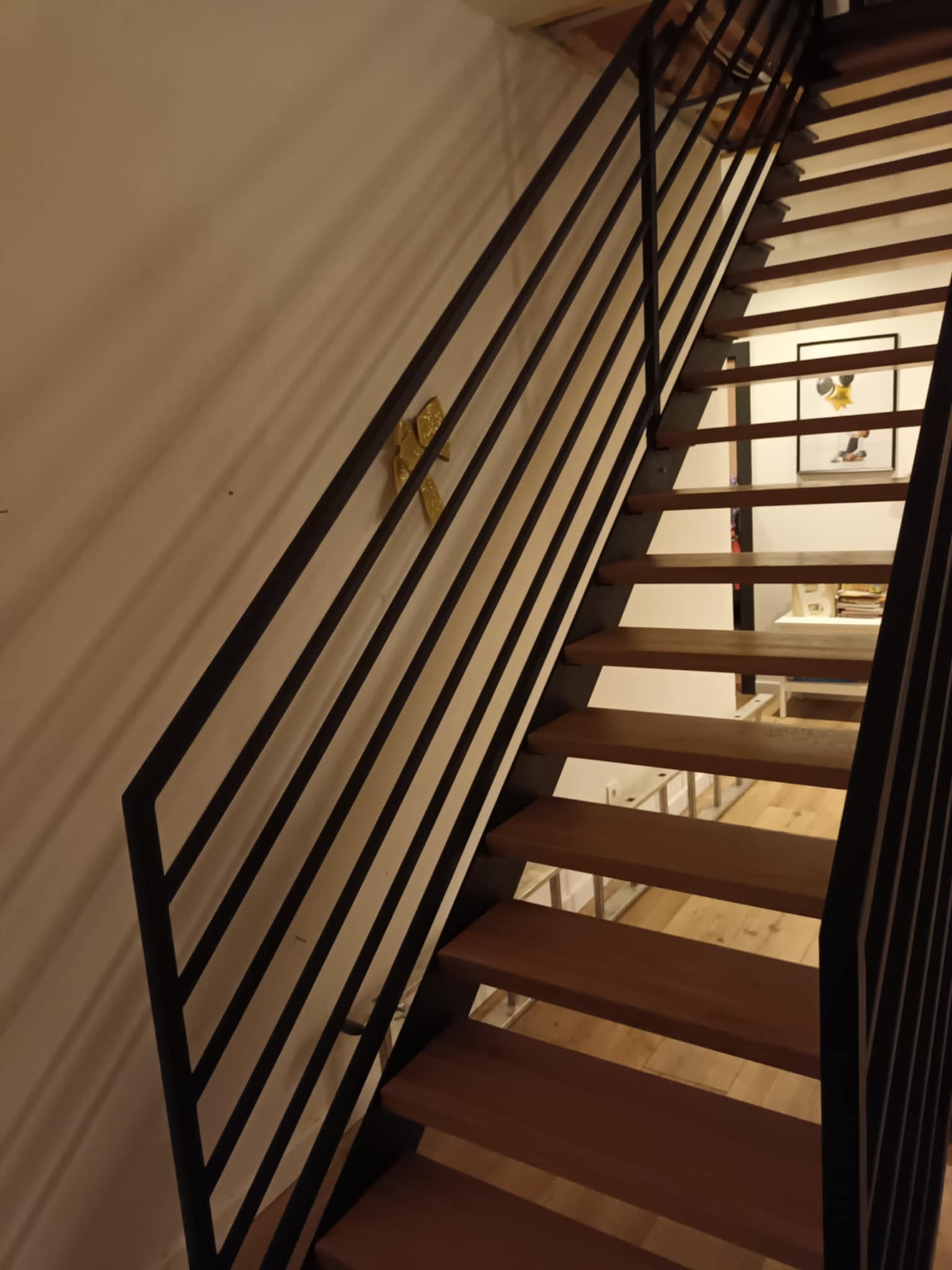 Nowoczesne schody drewniane z czarną, minimalistyczną balustradą z prętów; na ścianie widoczna złota ozdoba.