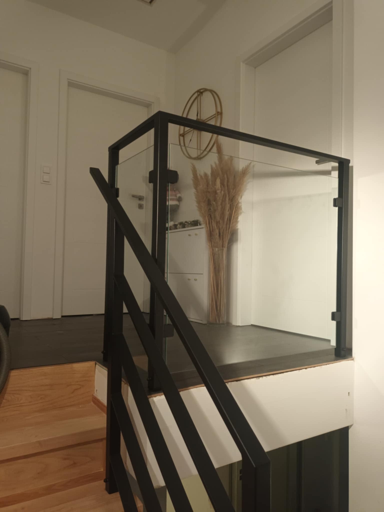 Szklana balustrada z czarnymi, metalowymi słupkami na schodach w jasnym wnętrzu. W tle widoczny dekoracyjny bukiet z suszonych traw i złoty zegar.