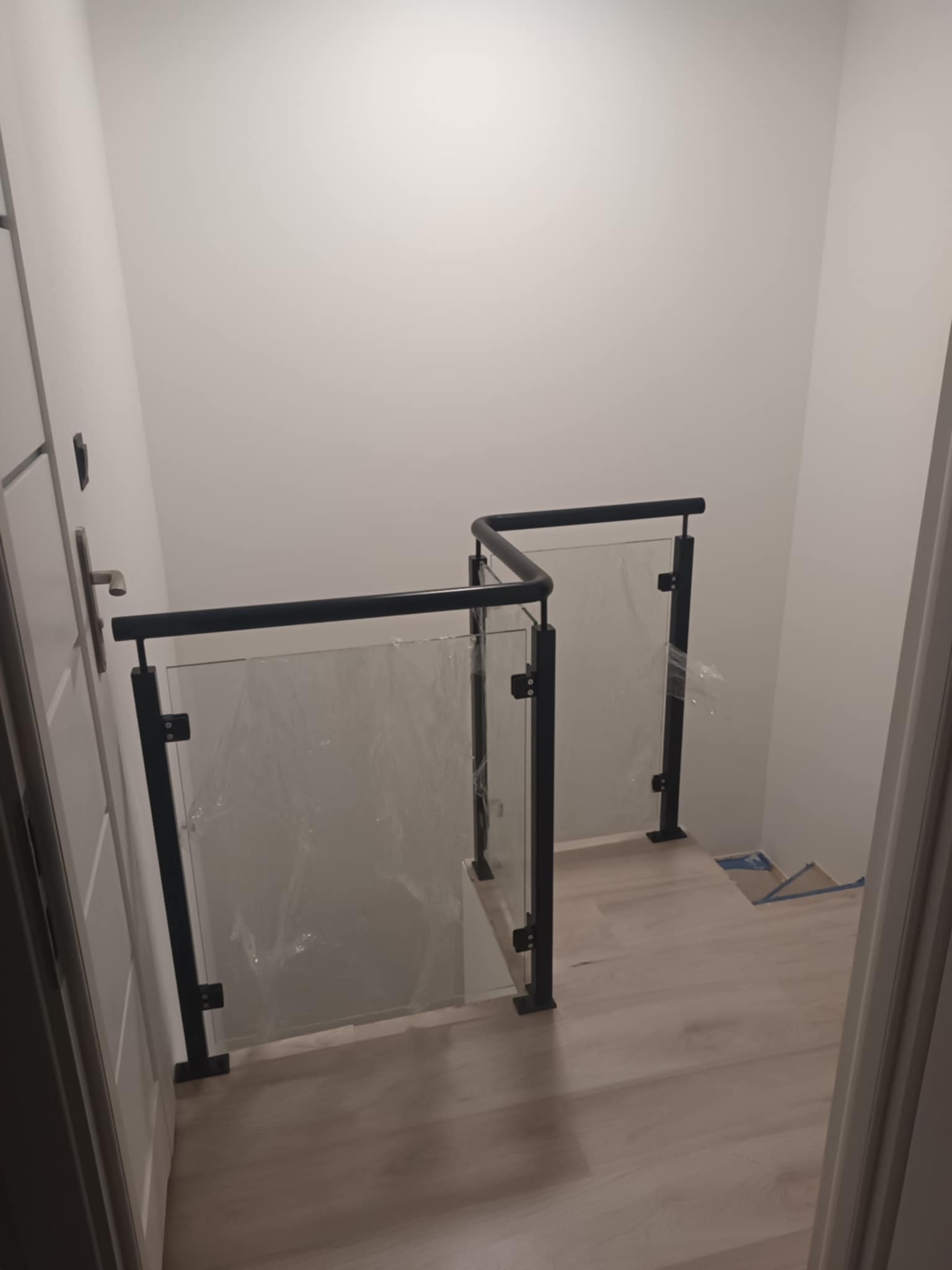 Szklana balustrada z czarnymi elementami mocującymi, zabezpieczona folią ochronną, zamontowana na schodach wewnątrz budynku, widok z góry.