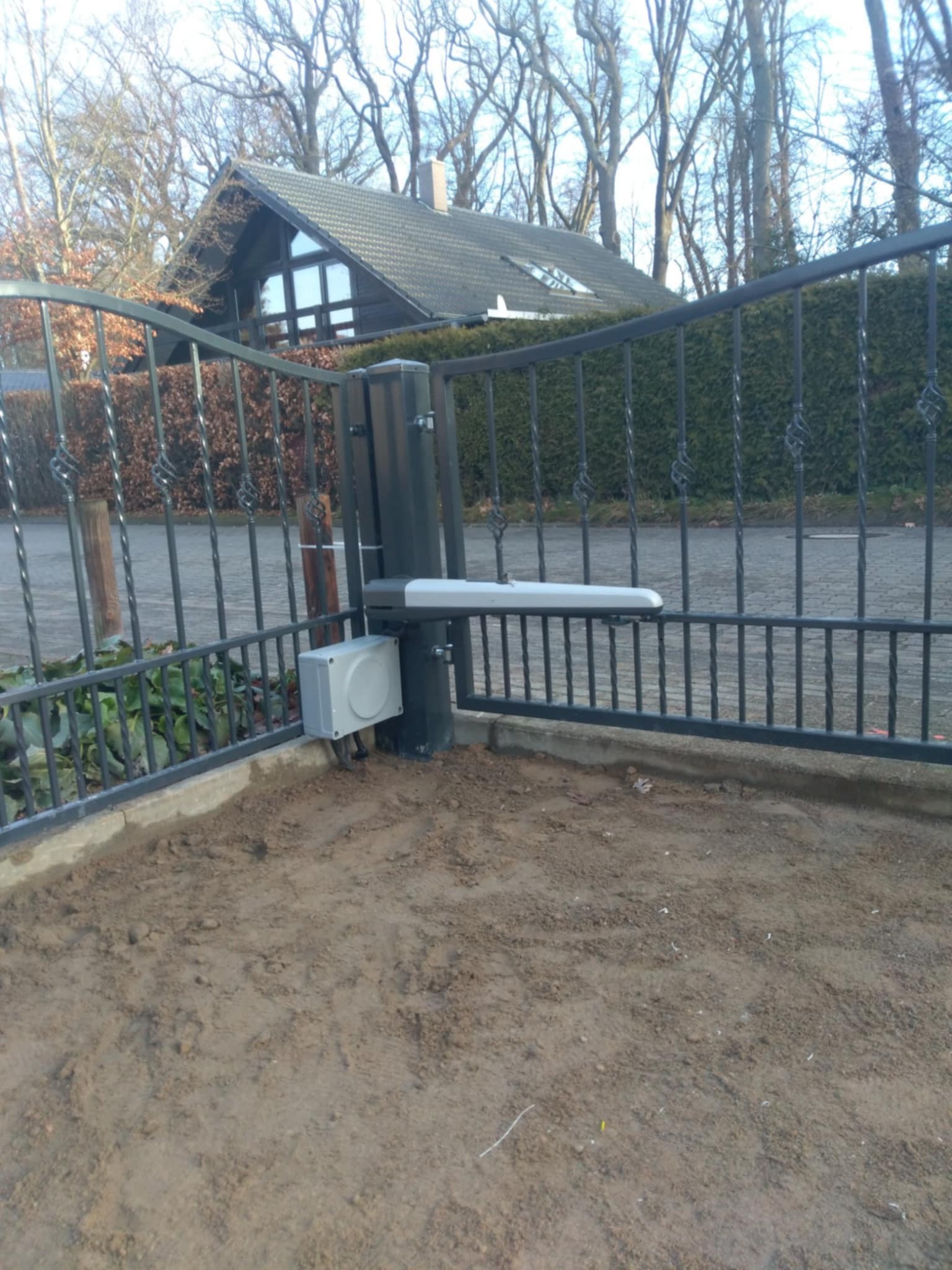 Czarna, metalowa brama wjazdowa z automatycznym napędem, widoczna skrzynka sterująca i ramię siłownika, w tle dom z ciemnym dachem i drzewa.