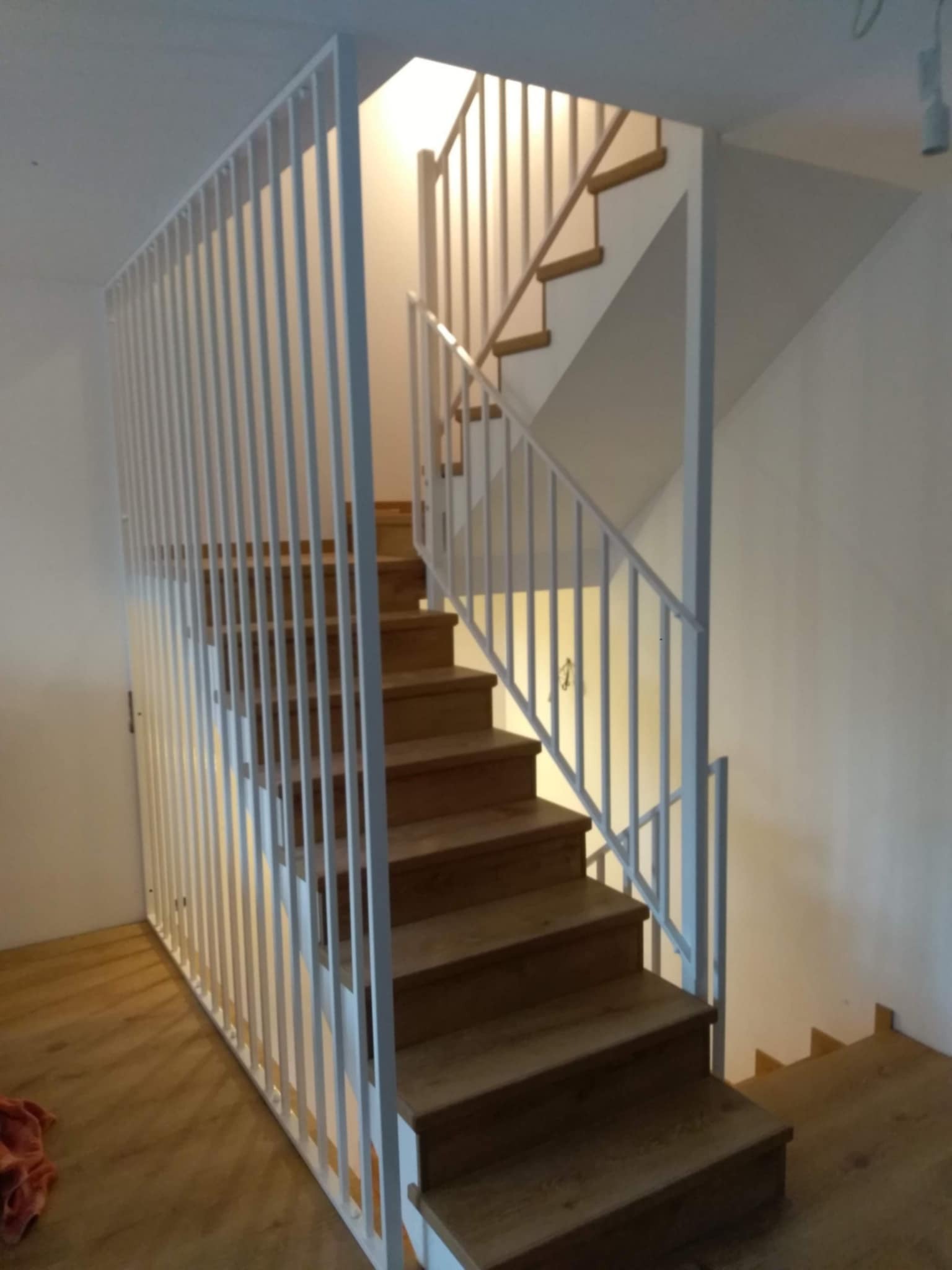 Drewniane schody z białą, pionową balustradą z metalowych prętów, widok z dołu, oświetlenie górne.