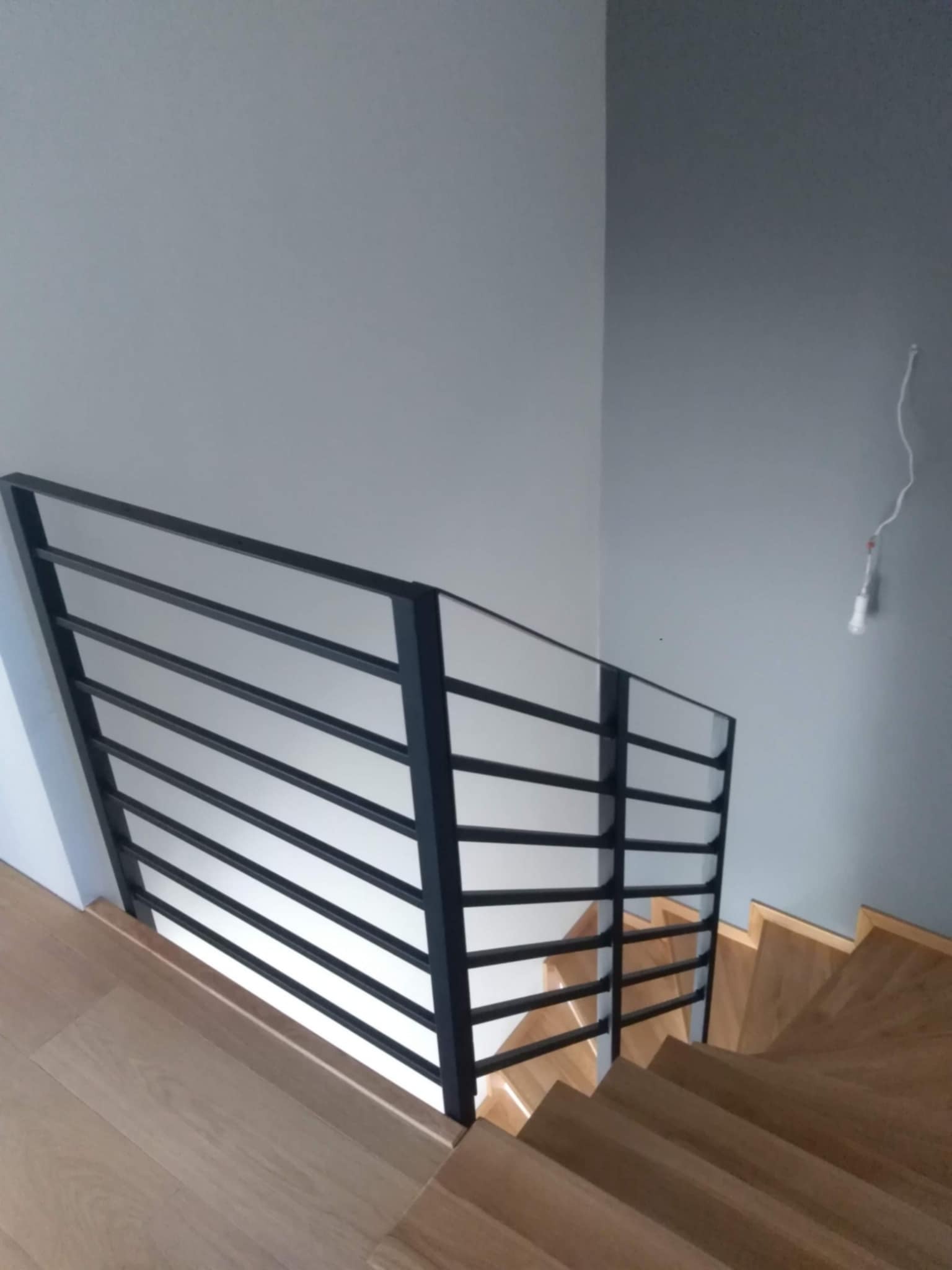 Minimalistyczna, czarna balustrada z poziomych prętów zamontowana przy drewnianych schodach w jasnym wnętrzu, widoczne niedokończone podłączenie elektryczne na ścianie.