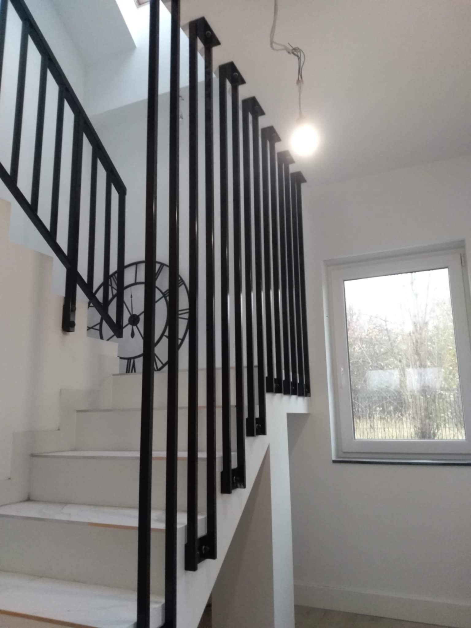 Białe schody z czarną, minimalistyczną balustradą, na ścianie dekoracyjny zegar, widok na zewnątrz przez okno.