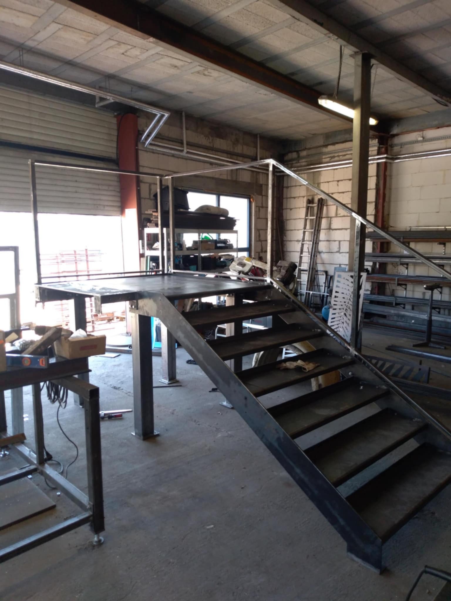 Konstrukcja schodów metalowych z platformą roboczą w warsztacie, widoczne narzędzia i elementy spawane, surowy stan przed wykończeniem.