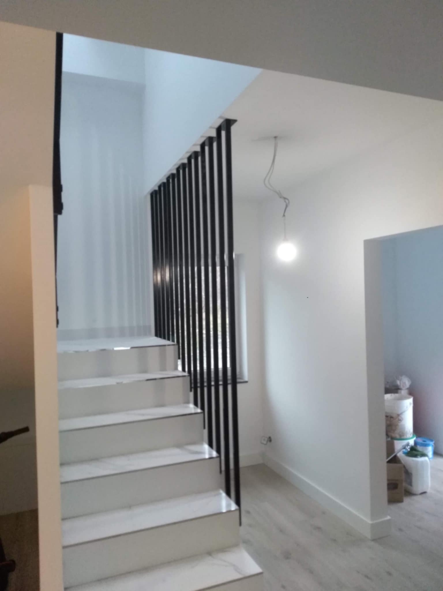 Wnętrze domu z białymi schodami wykończonymi imitacją marmuru i czarną, pionową balustradą z metalowych prętów, widok z dołu na górę, lampa zwisająca na kablu.