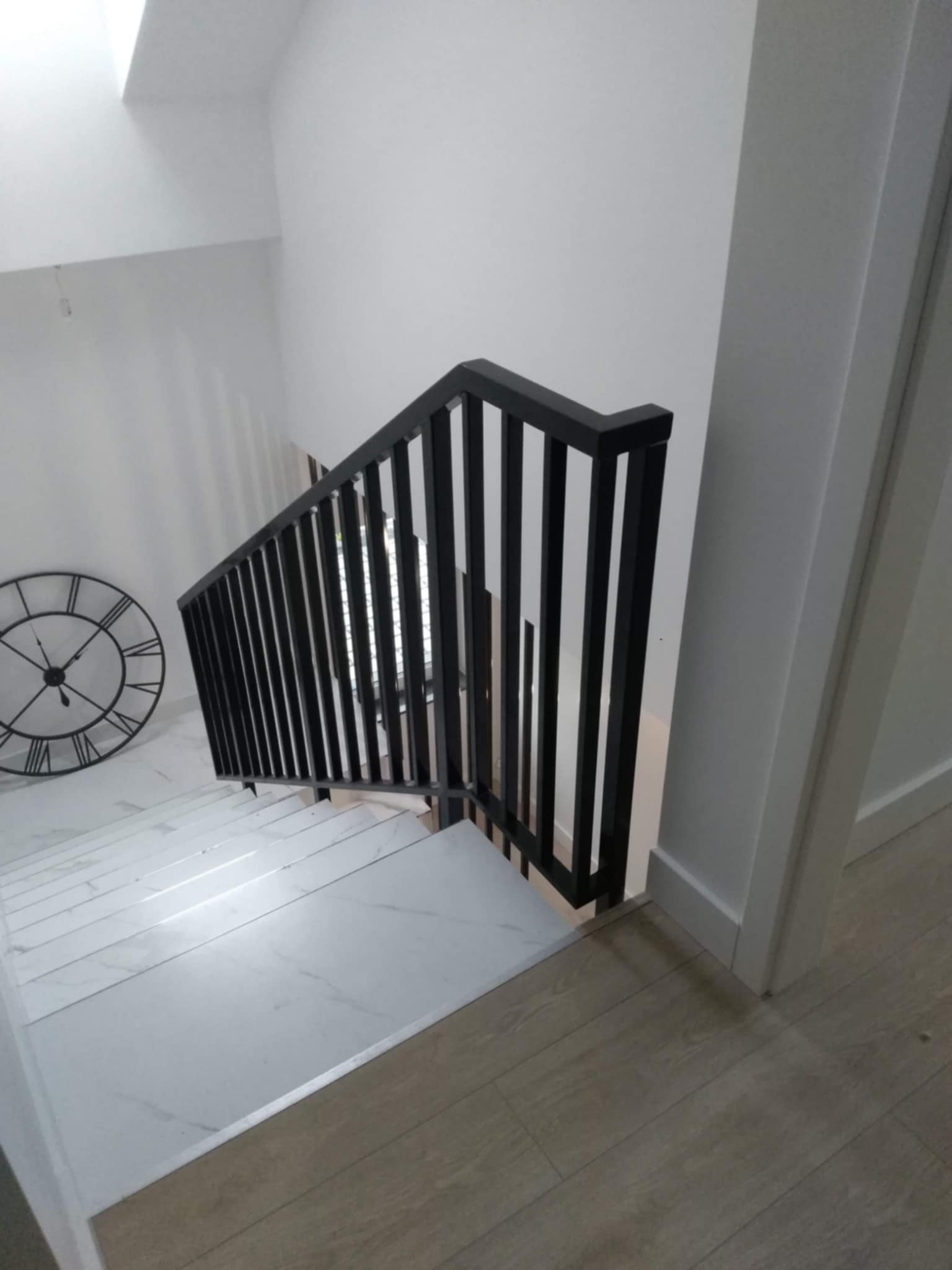 Nowoczesna, czarna balustrada schodowa z pionowymi szczeblami, na białych marmurowych schodach, widok z góry, z dekoracyjnym zegarem na ścianie.