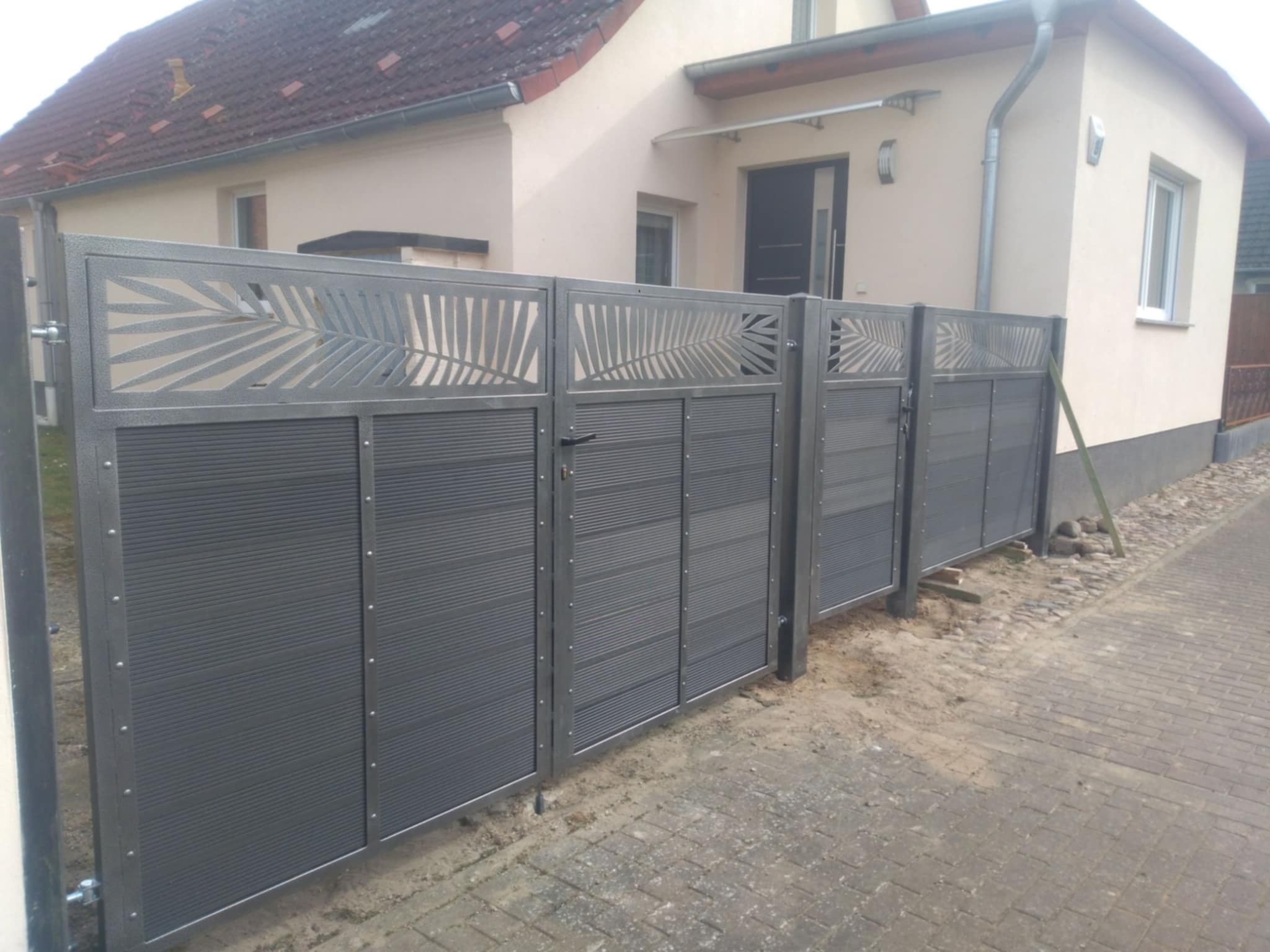 Nowoczesne ogrodzenie panelowe w kolorze grafitowym z dekoracyjnymi wycięciami w kształcie liści palmowych, zamontowane przed domem z beżową elewacją i brązowym dachem.