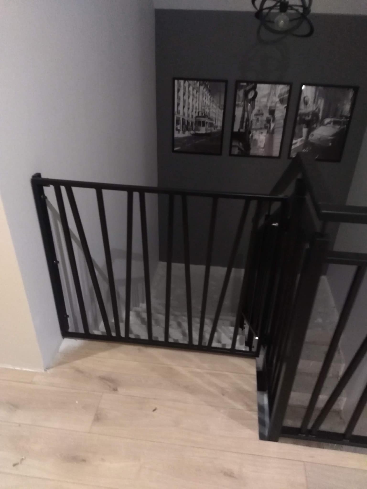 Nowoczesna, czarna barierka schodowa z ukośnymi prętami, zamontowana wewnątrz domu na jasnej podłodze, z widocznymi schodami i trzema czarno-białymi obrazami na szarej ścianie w tle.
