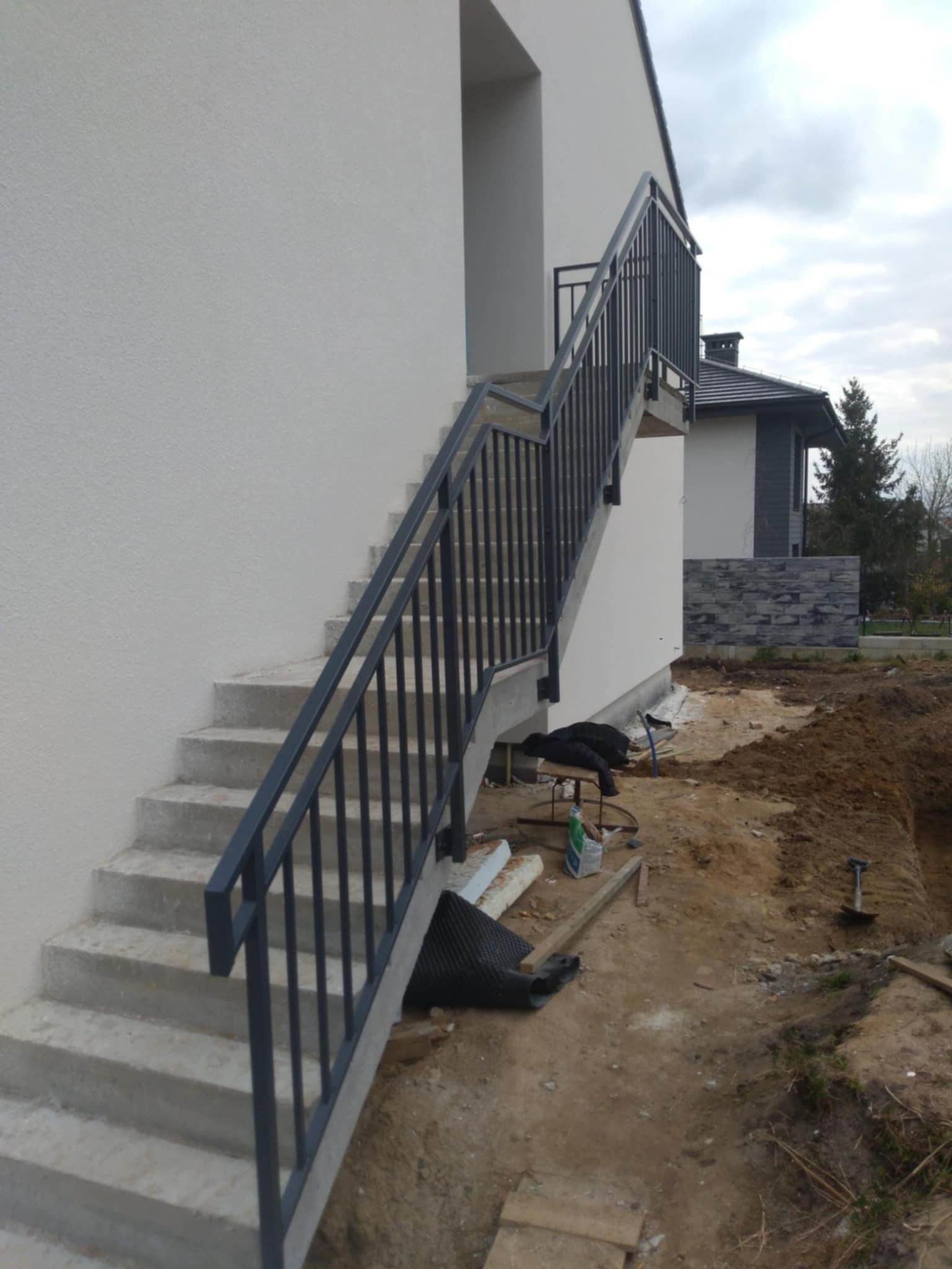 Betonowe schody zewnętrzne z czarną metalową balustradą przy nowo wybudowanym domu, teren wokół w trakcie zagospodarowania.