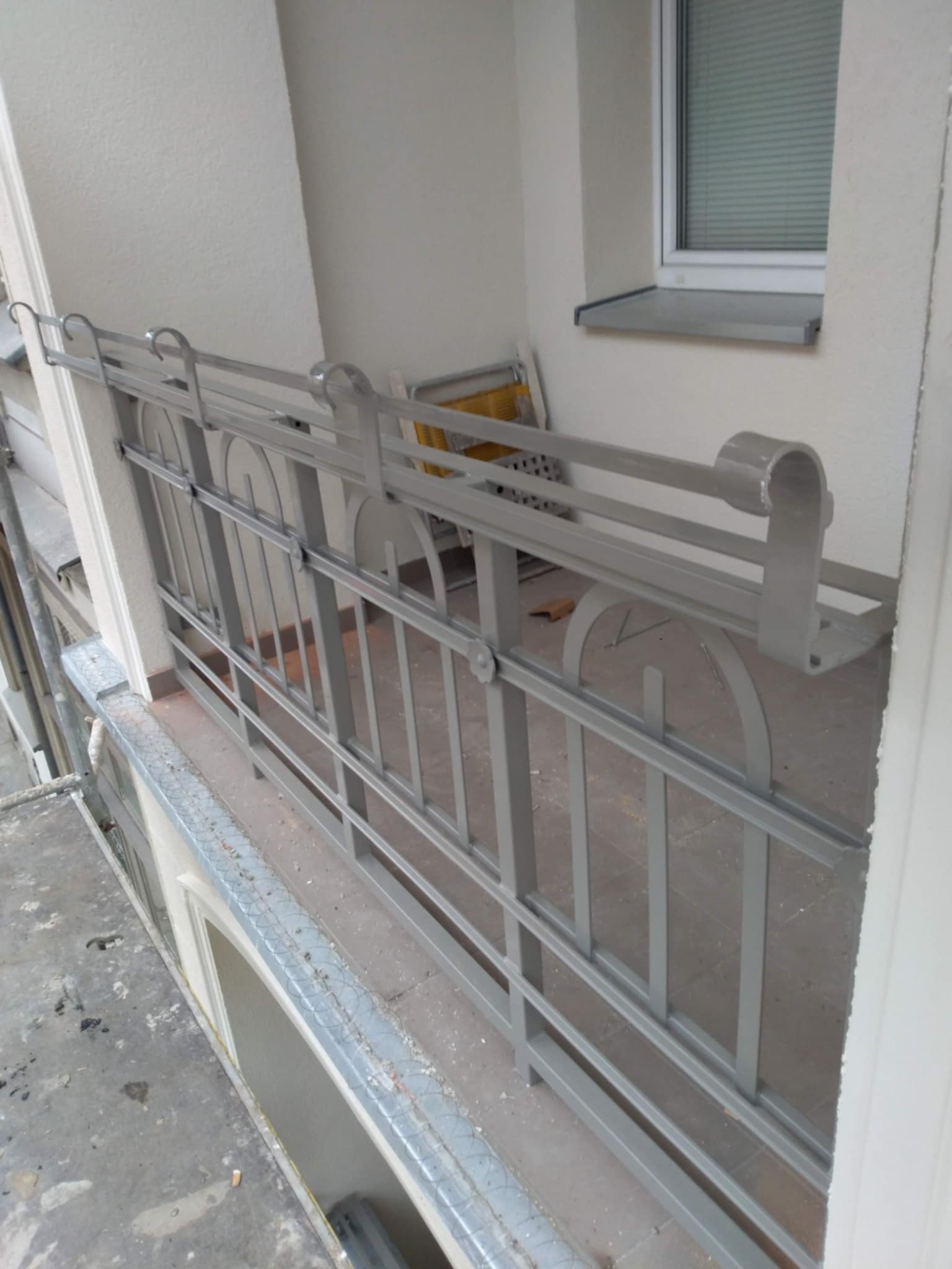 Szara metalowa balustrada balkonowa z dekoracyjnymi elementami w kształcie półkola, widoczna z bliska, z oknem w tle.