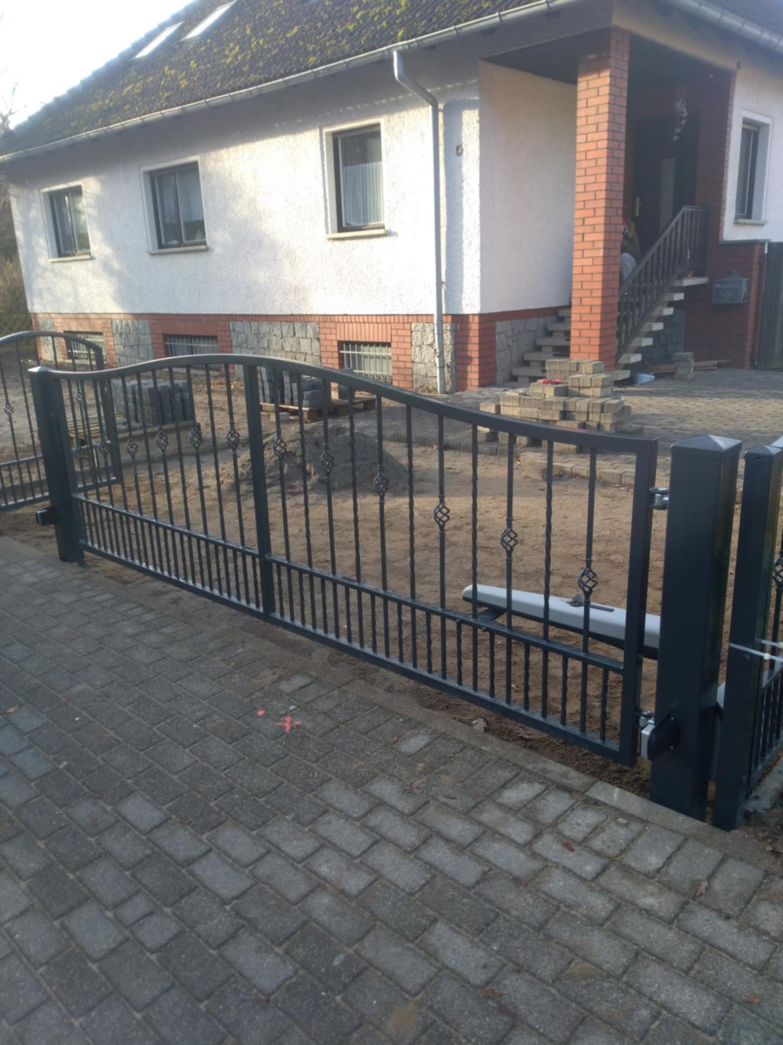 Automatyczna brama wjazdowa z czarnego metalu, z dekoracyjnymi elementami, zamontowana przed domem w trakcie budowy, widoczny mechanizm otwierający.