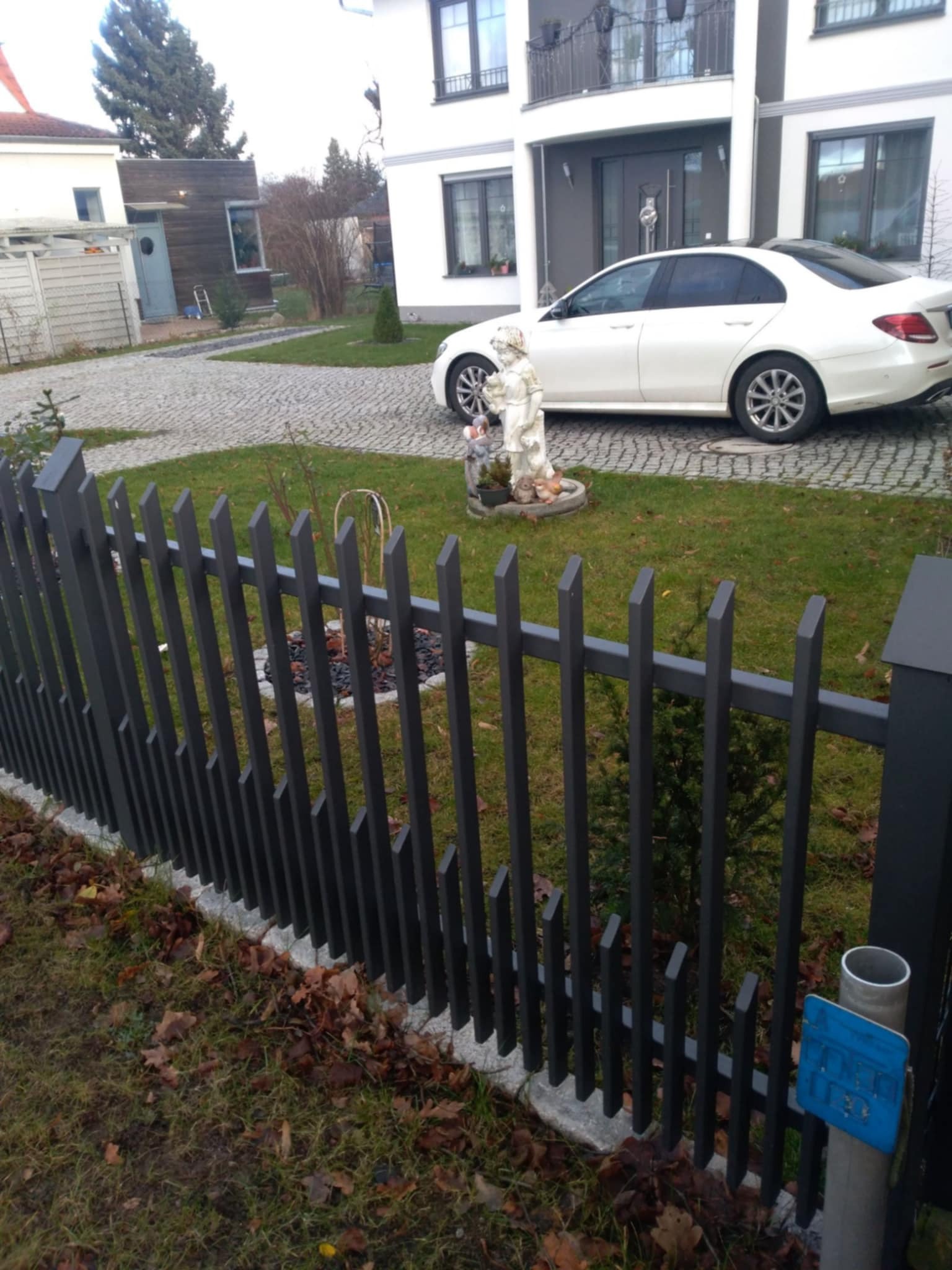 Nowoczesne ogrodzenie z metalowych sztachet o skośnych zakończeniach, widok na trawnik, rzeźbę ogrodową i biały samochód zaparkowany przed domem.