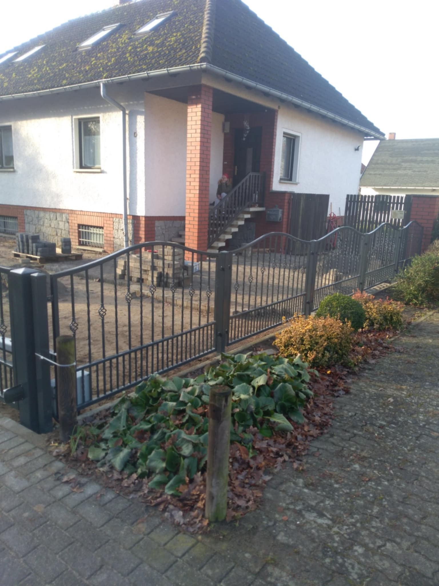 Metalowe ogrodzenie posesji z bramą wjazdową, widoczne stosy kostki brukowej przygotowane do dalszych prac wykończeniowych.