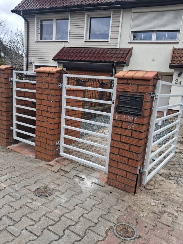 Biała, pozioma brama wjazdowa z ceglanymi słupkami zwieńczonymi dachówką, zamontowana przed domem z beżową elewacją i czerwoną dachówką. Widok z poziomu chodnika.