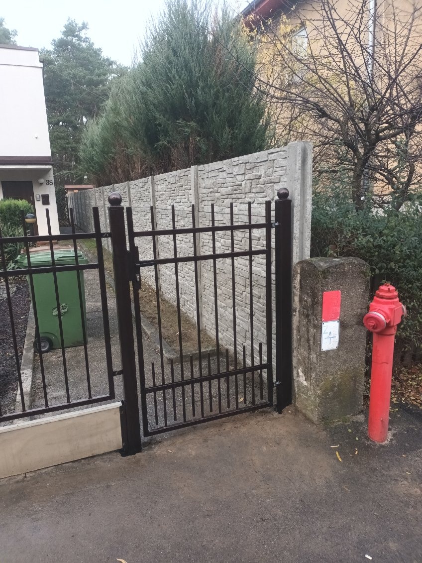 Ciemna, metalowa furtka z pionowymi prętami, zamykająca wejście na posesję. W tle betonowy mur z fakturą kamienia i zielone tuje. Po prawej hydrant.
