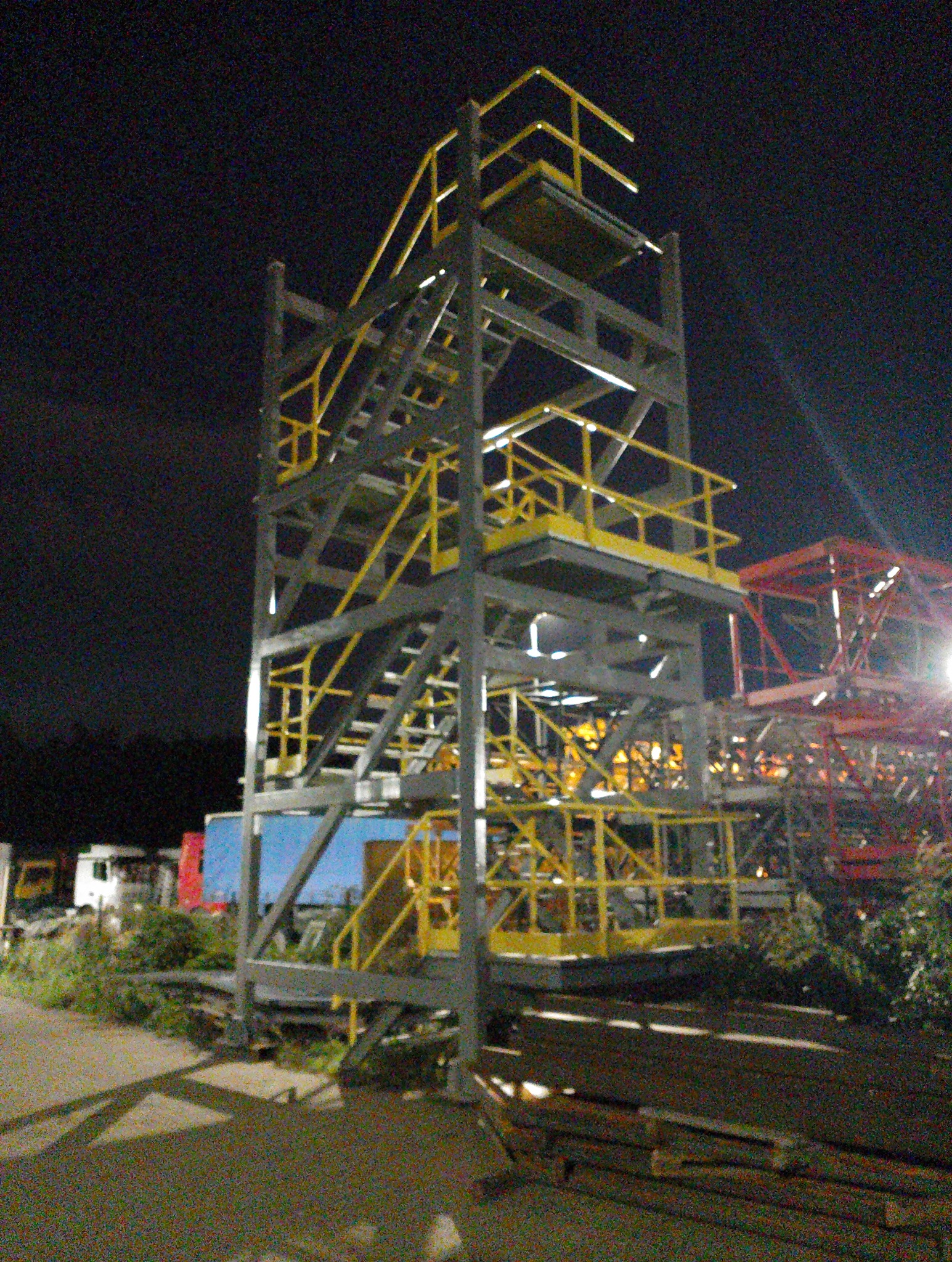 Konstrukcja stalowa z żółtymi poręczami i schodami, oświetlona nocą. Widoczne elementy rusztowania i inne konstrukcje w tle. Niska jakość zdjęcia.