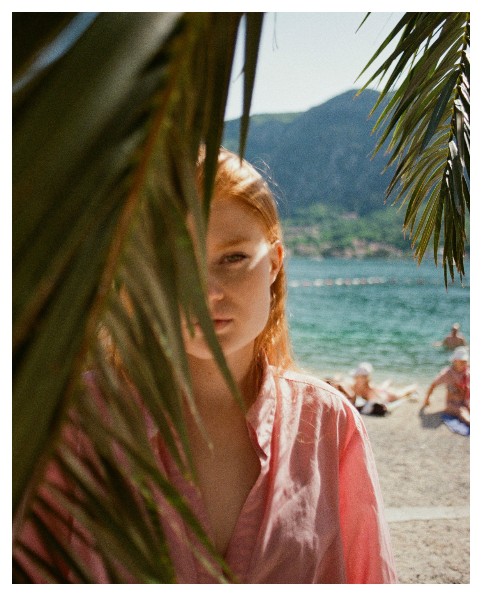 Portret kobiety o rudych włosach w różowej koszuli, częściowo zasłoniętej liśćmi palmy, z plażą i morzem w tle.
