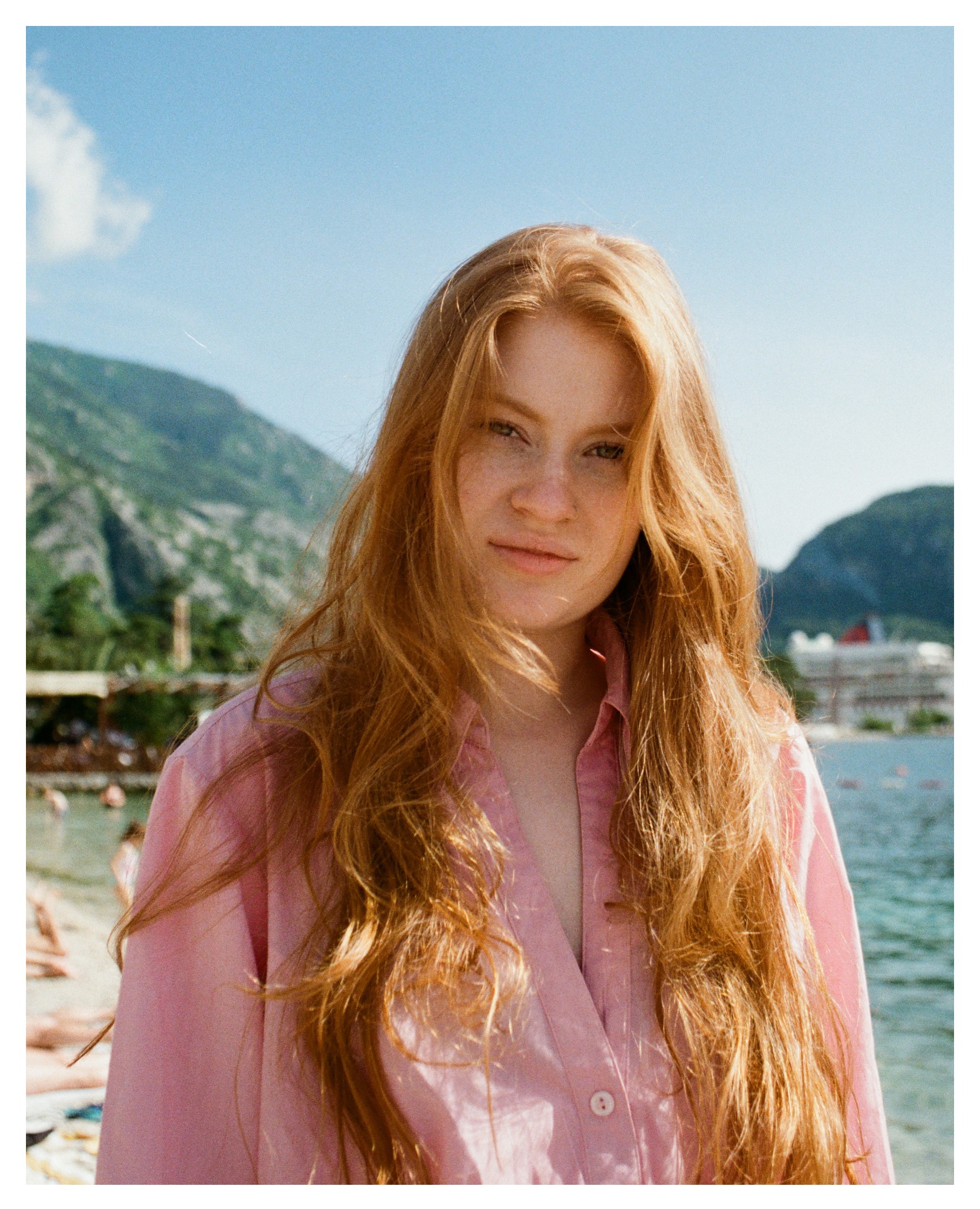 Portret rudej kobiety w różowej koszuli na tle plaży i gór, wykonany w stylu vintage z widocznym ziarnem.