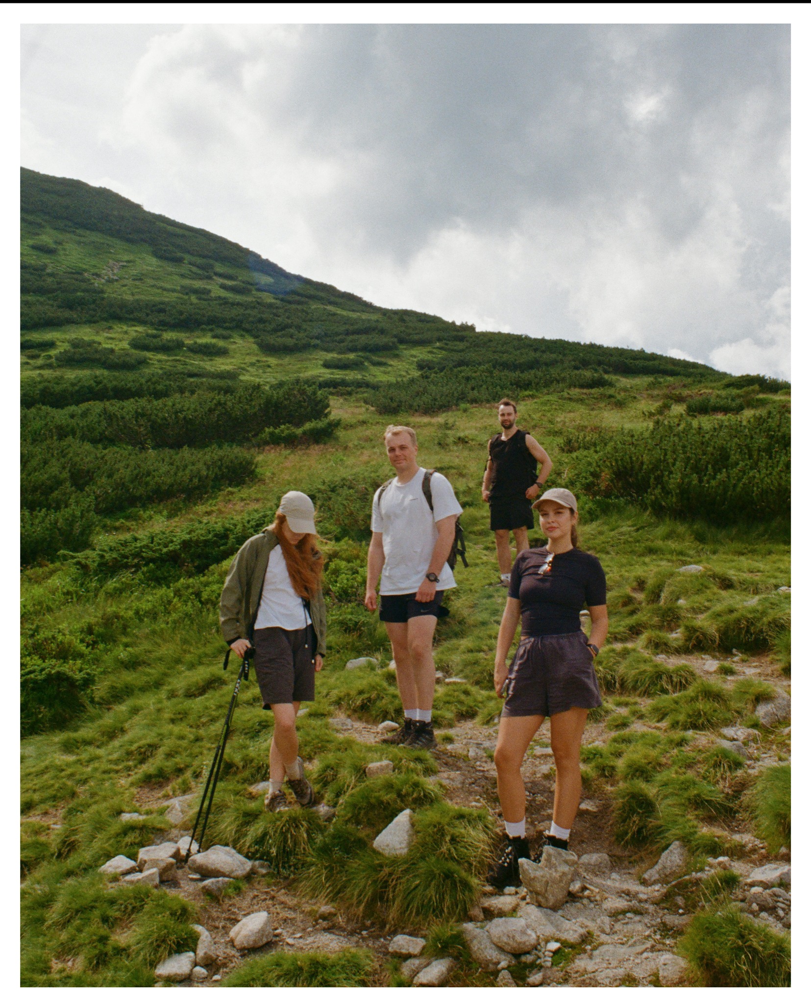 Grupa czterech osób podczas wędrówki po górskim szlaku; trawiasta zbocze, kamienie, pochmurne niebo.