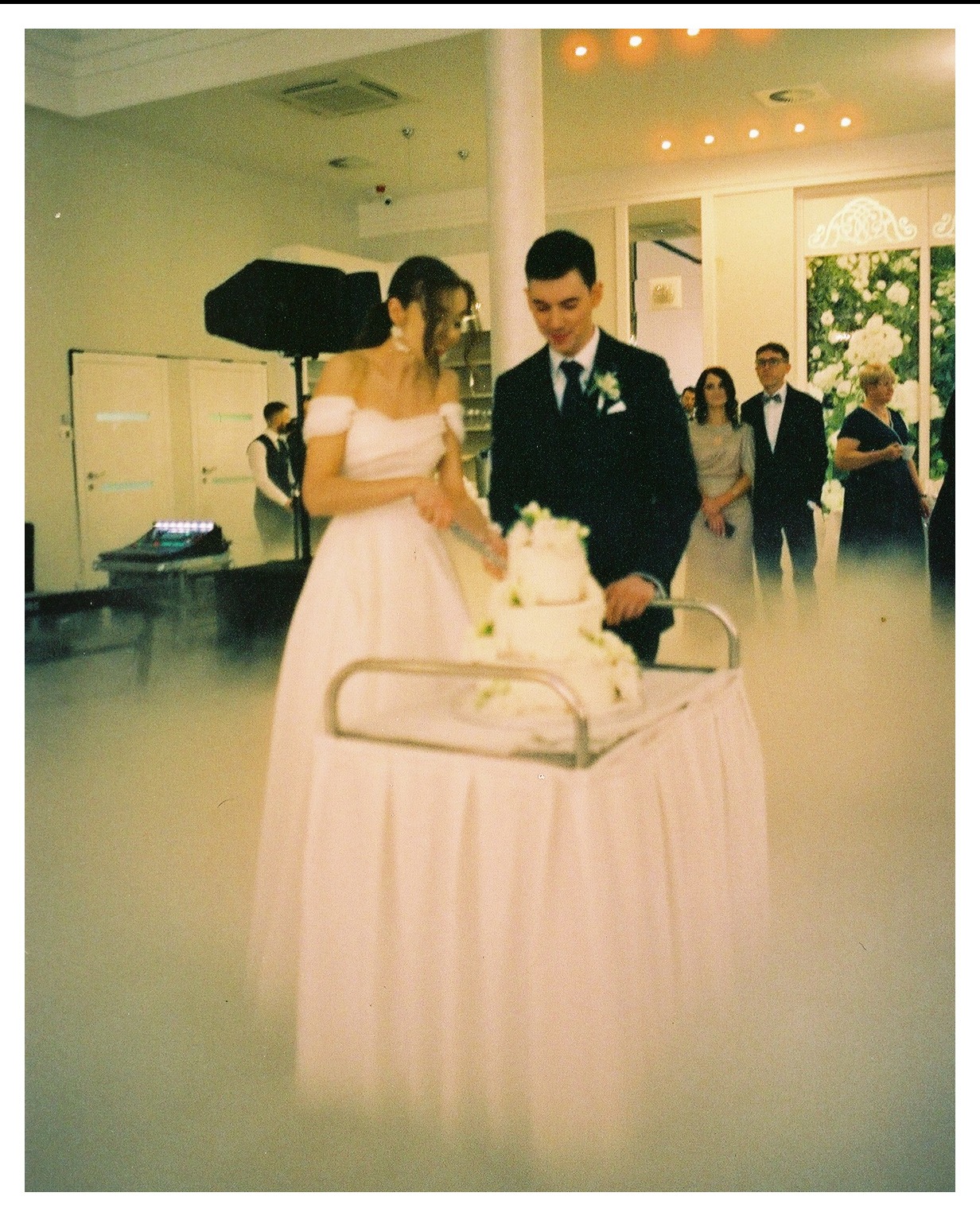 Para młoda kroi tort weselny na stole otoczonym suchym lodem, w tle goście i oświetlenie sceniczne.