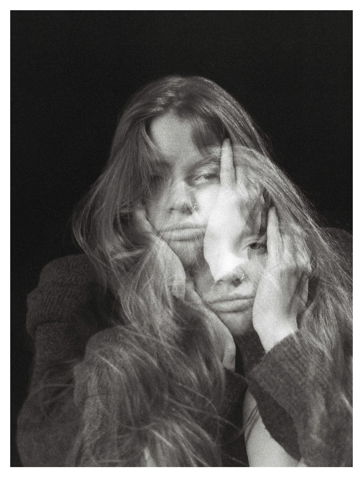 Czarno-białe, artystyczne zdjęcie kobiety z długimi włosami, z efektem podwójnej ekspozycji, opierającej twarz na dłoniach, z kolczykiem w nosie, na ciemnym tle.