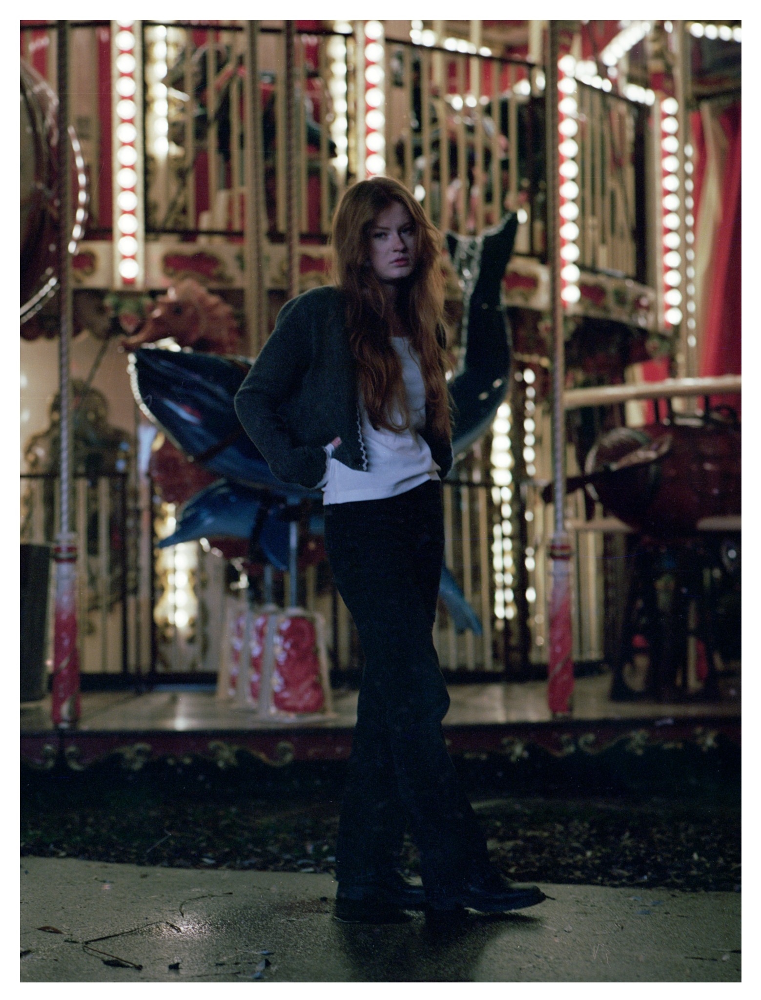 Młoda kobieta o rudych włosach pozuje na tle karuzeli z podświetleniem, ubrana w ciemne spodnie, białą koszulkę i szary sweter