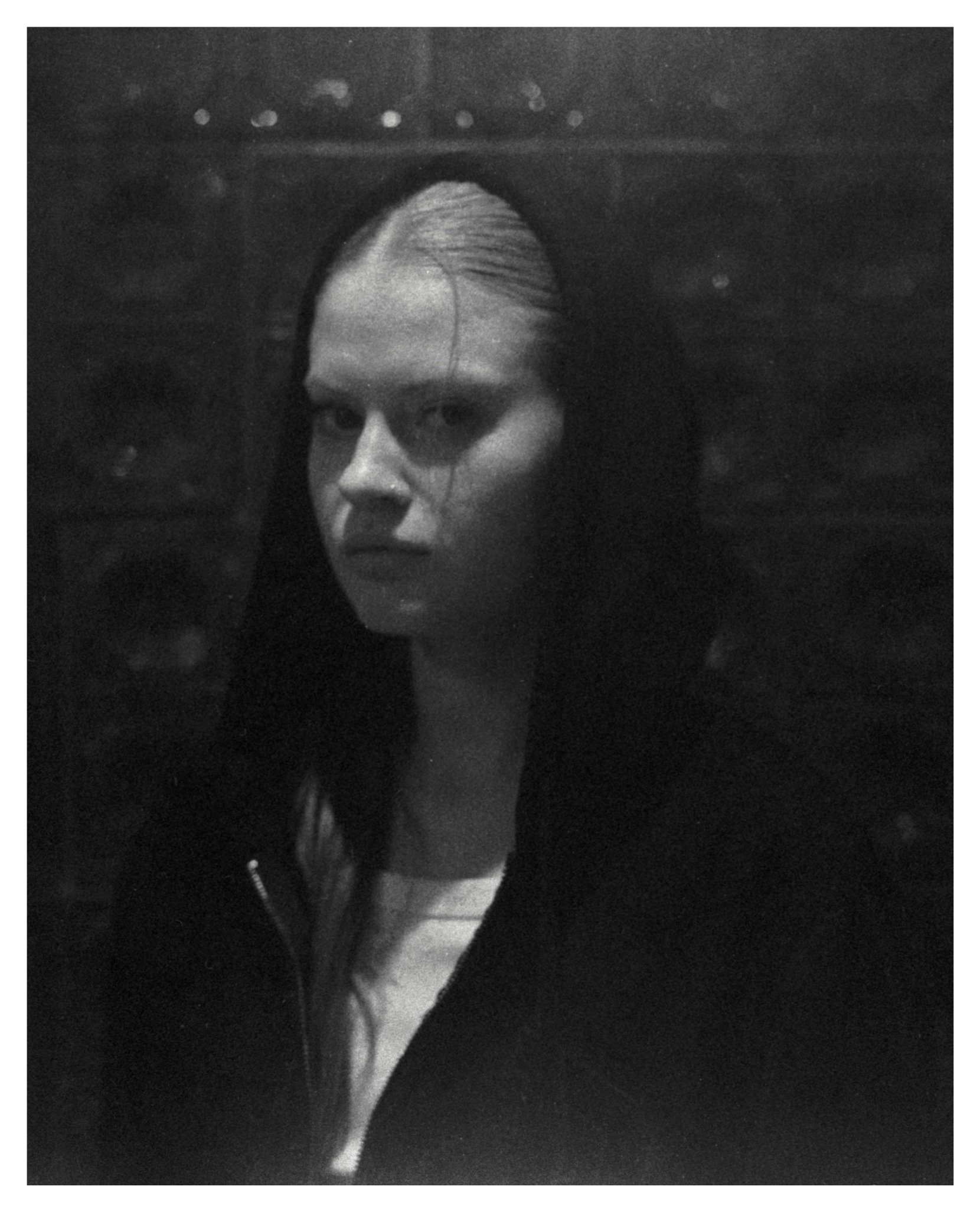 Czarno-biały portret kobiety w kapturze z zaciętym wyrazem twarzy, oświetlonej od góry, z widoczną fakturą ziarna fotograficznego.