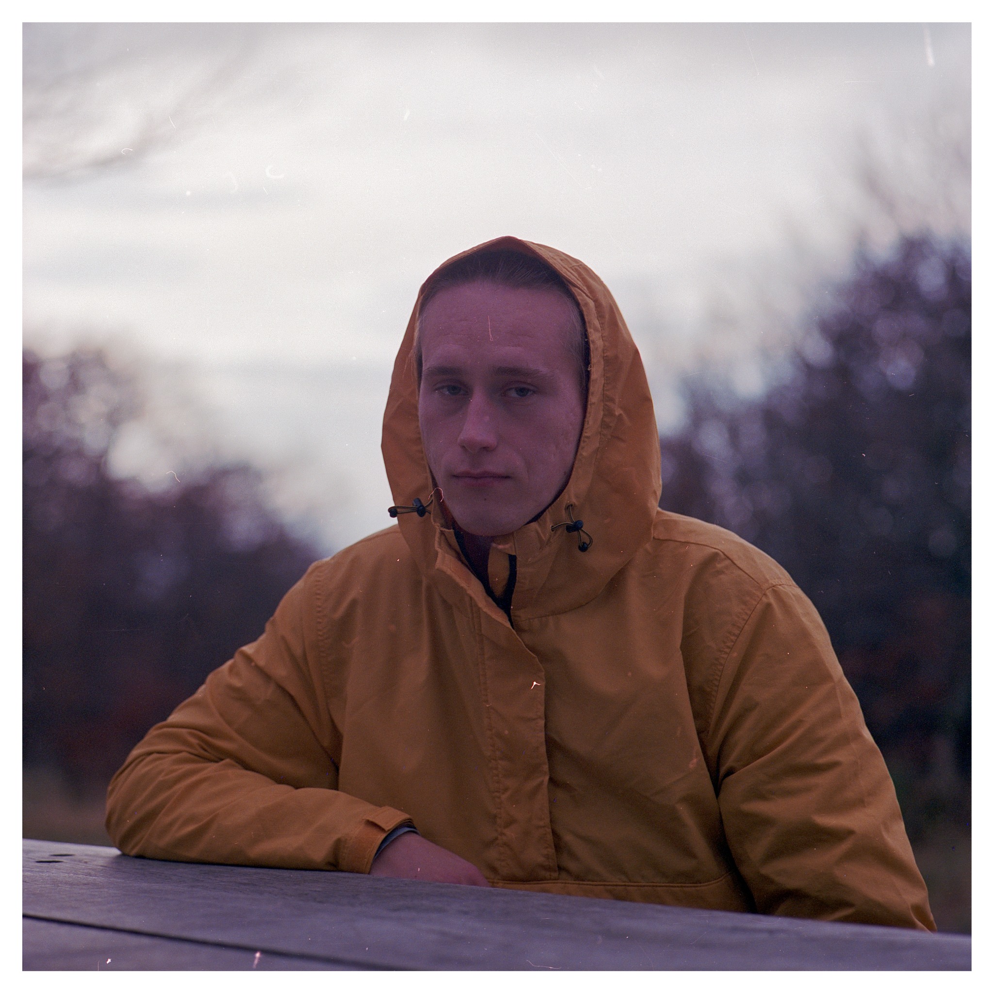 Portret młodego mężczyzny w żółtej kurtce z kapturem, siedzącego przy drewnianym stole na zewnątrz, z rozmazanym tłem drzew i pochmurnego nieba.