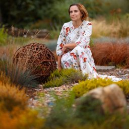 Miroslaw Kocurek Photography - Kobieta w kwiecistej sukience klęczy w ogrodzie obok dekoracyjnej kuli z gałęzi, spoglądając w dal.