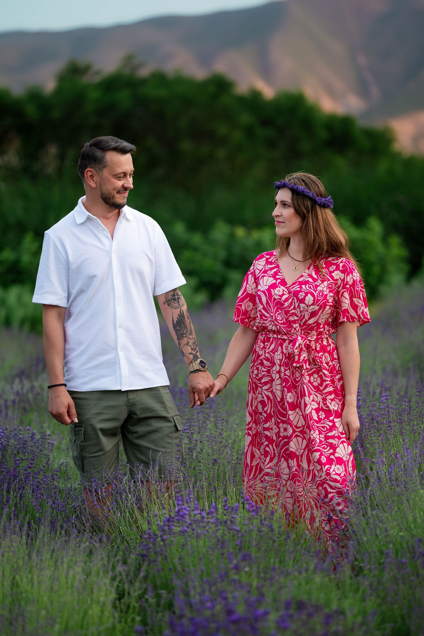 Para trzymająca się za ręce na polu lawendy, kobieta w kwiecistej sukience i wianku, mężczyzna z tatuażem na ramieniu, w tle zieleń drzew.