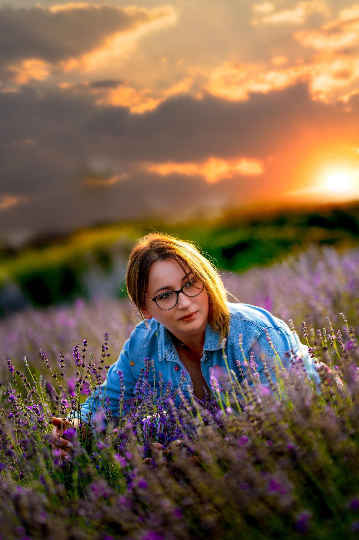 Kobieta w okularach, w denimowej koszuli, delikatnie dotyka kwiatów lawendy na polu w Rzeszowie, w tle zachodzące słońce.