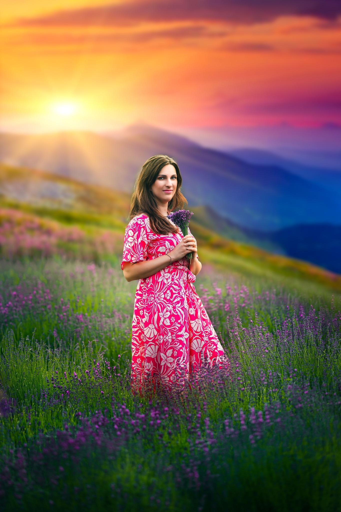 Kobieta w kwiecistej sukience trzyma bukiet lawendy na tle górskiego krajobrazu o zachodzie słońca.