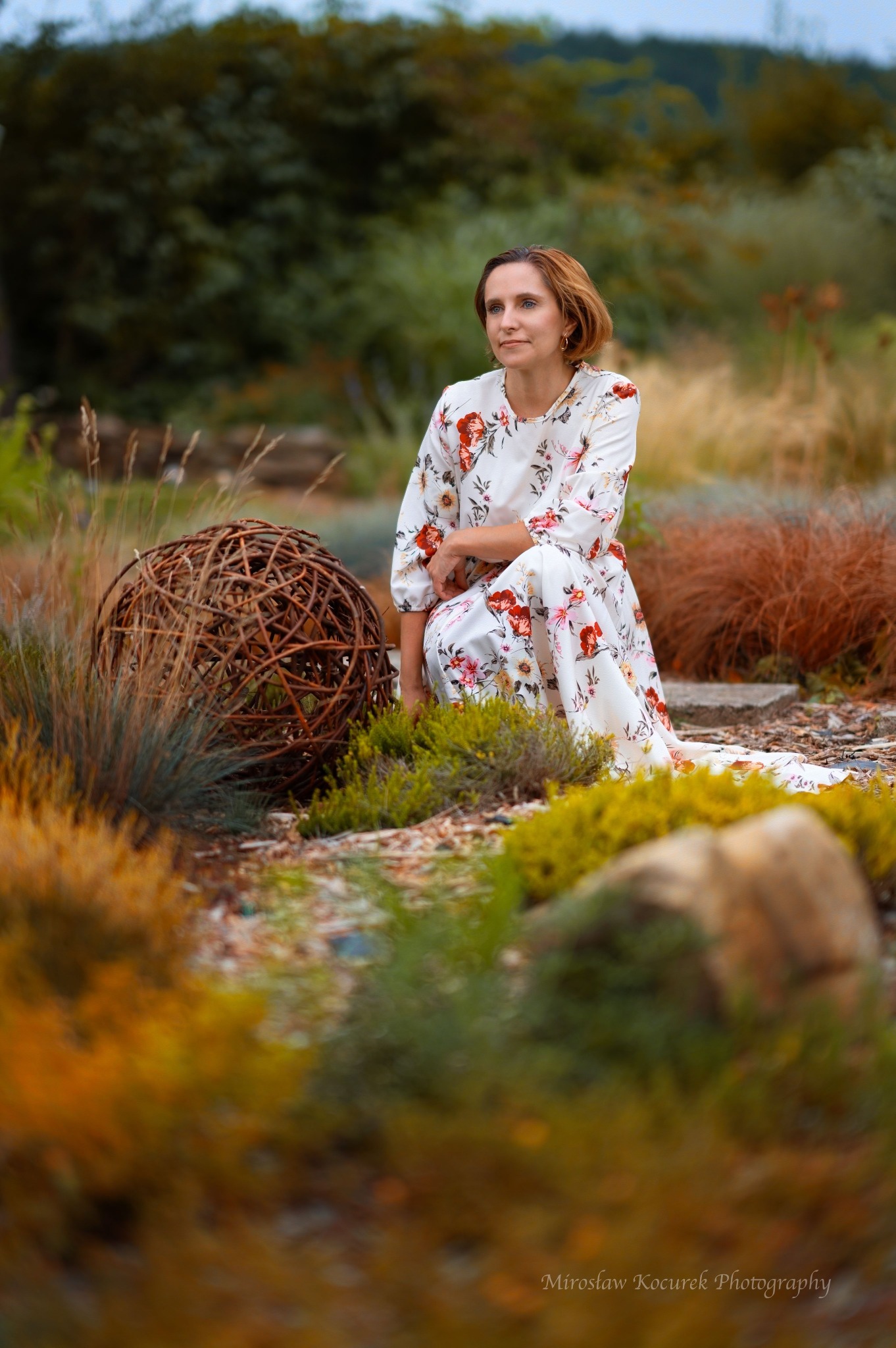 Kobieta w kwiecistej sukience klęczy w ogrodzie obok dekoracyjnej kuli z gałęzi, spoglądając w dal.