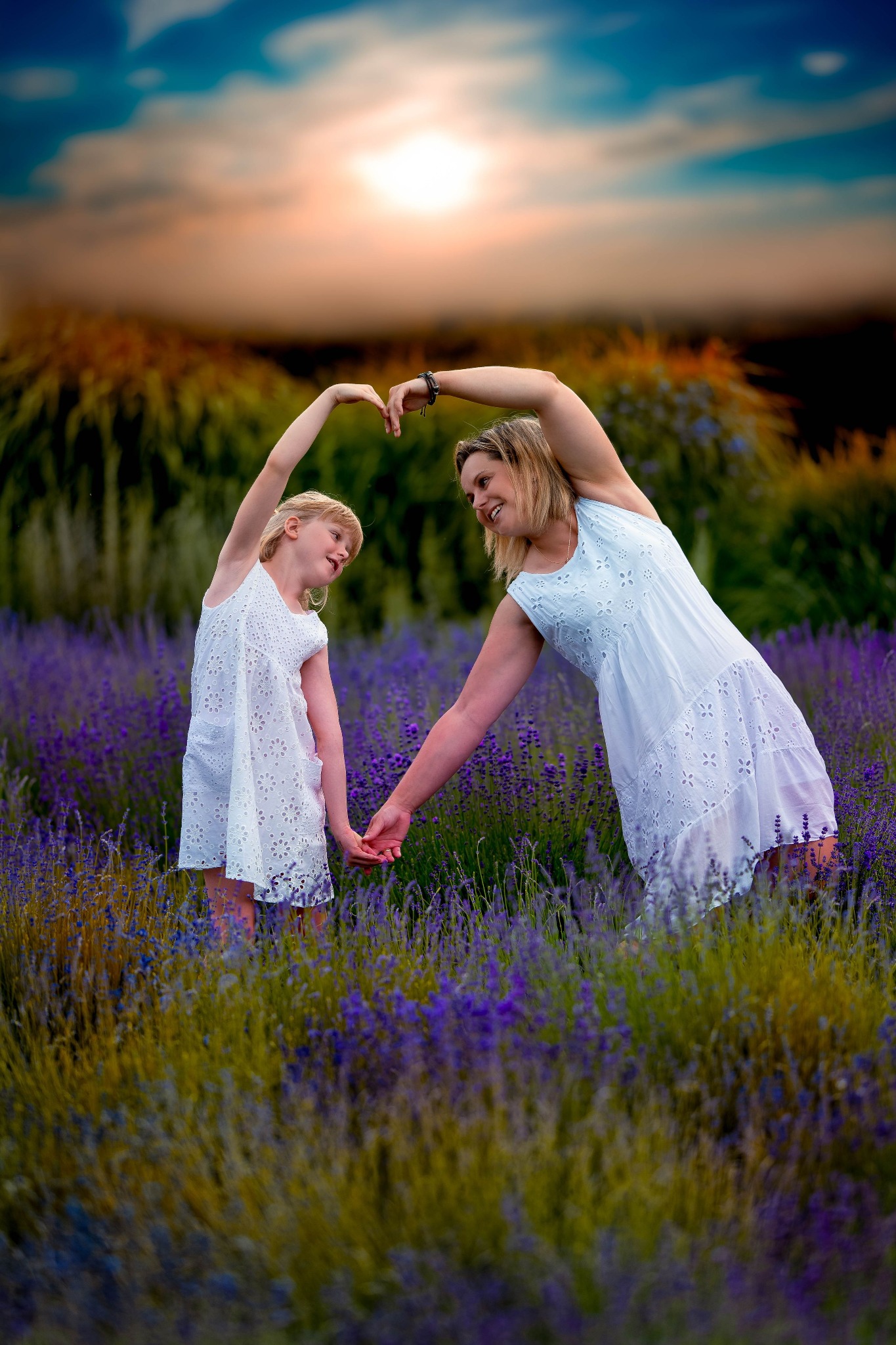Matka i córka w białych sukienkach stoją na polu lawendy, trzymają się za ręce i tworzą kształt serca z ramion nad głowami, zachód słońca w tle.