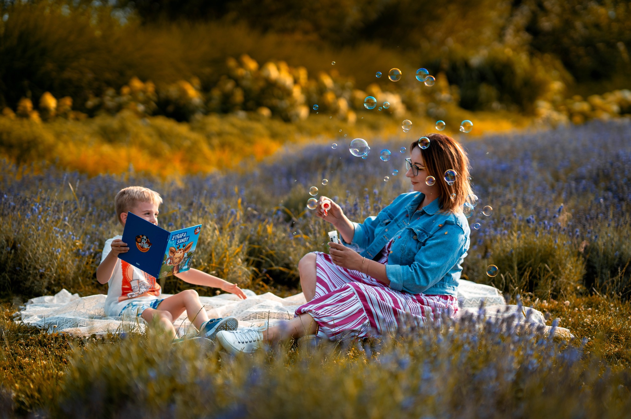 Kobieta w dżinsowej kurtce puszcza bańki mydlane dla chłopca czytającego książkę na kocu w kwitnącym polu lawendy, ciepłe światło słoneczne.