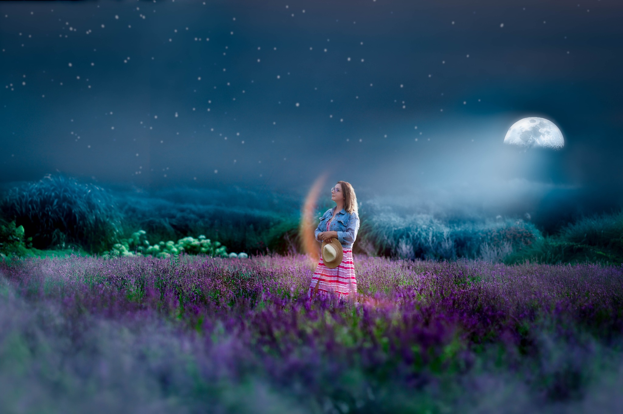 Kobieta w sukience w paski i dżinsowej kurtce stoi na fioletowym polu lawendy pod rozgwieżdżonym nocnym niebem z księżycem, trzymając słomkowy kapelusz.