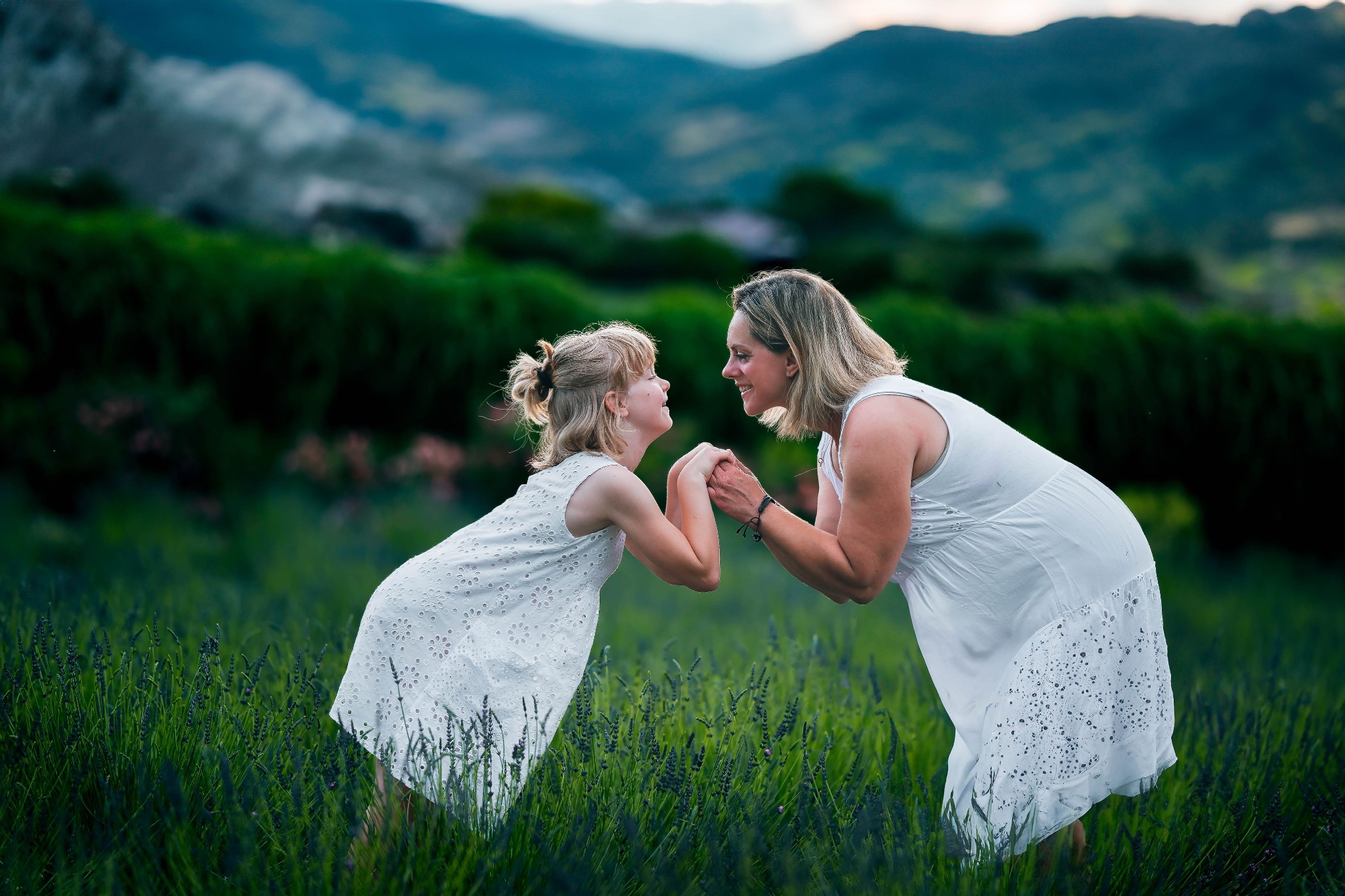Matka i córka w białych sukienkach stoją naprzeciw siebie na polu lawendy, trzymając się za ręce i uśmiechając się, z górami w tle.