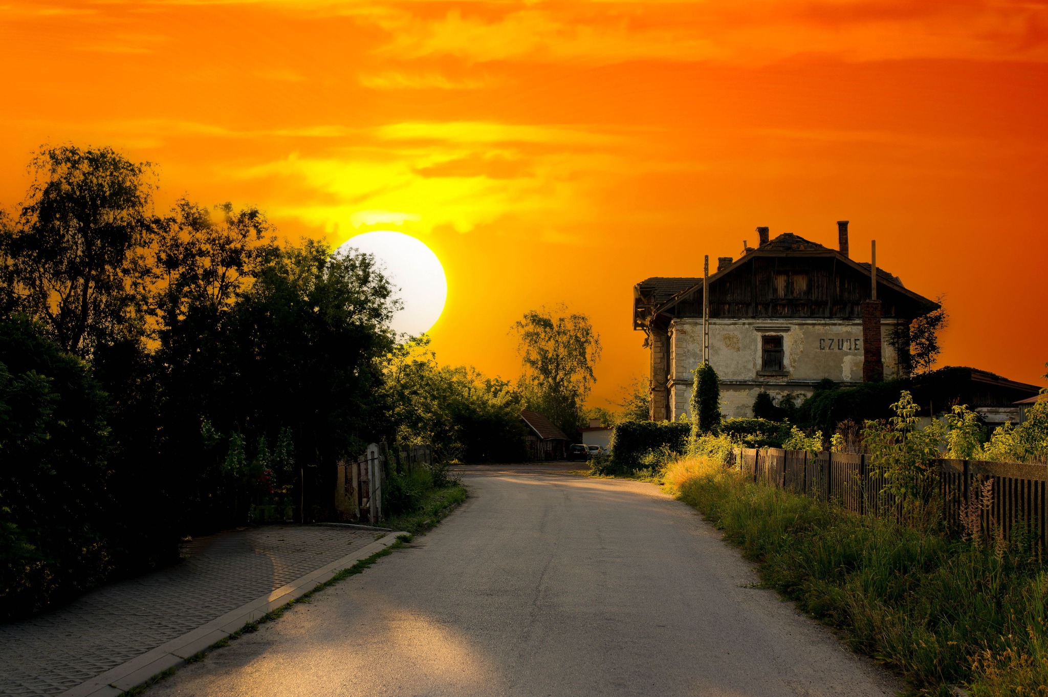 Droga prowadząca do starego, zaniedbanego budynku z napisem 'CZUJE' pod zachodzącym słońcem, otoczona zielenią i drewnianym płotem.