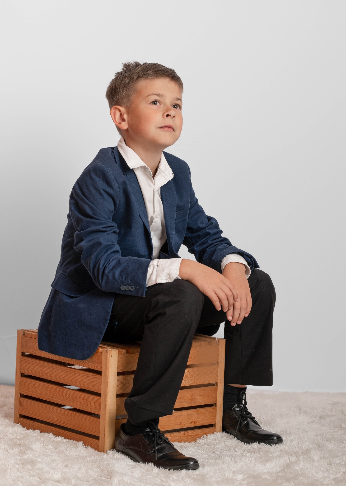 Chłopiec w granatowej marynarce i czarnych spodniach siedzi na drewnianej skrzynce, spoglądając w górę na jasnym tle.