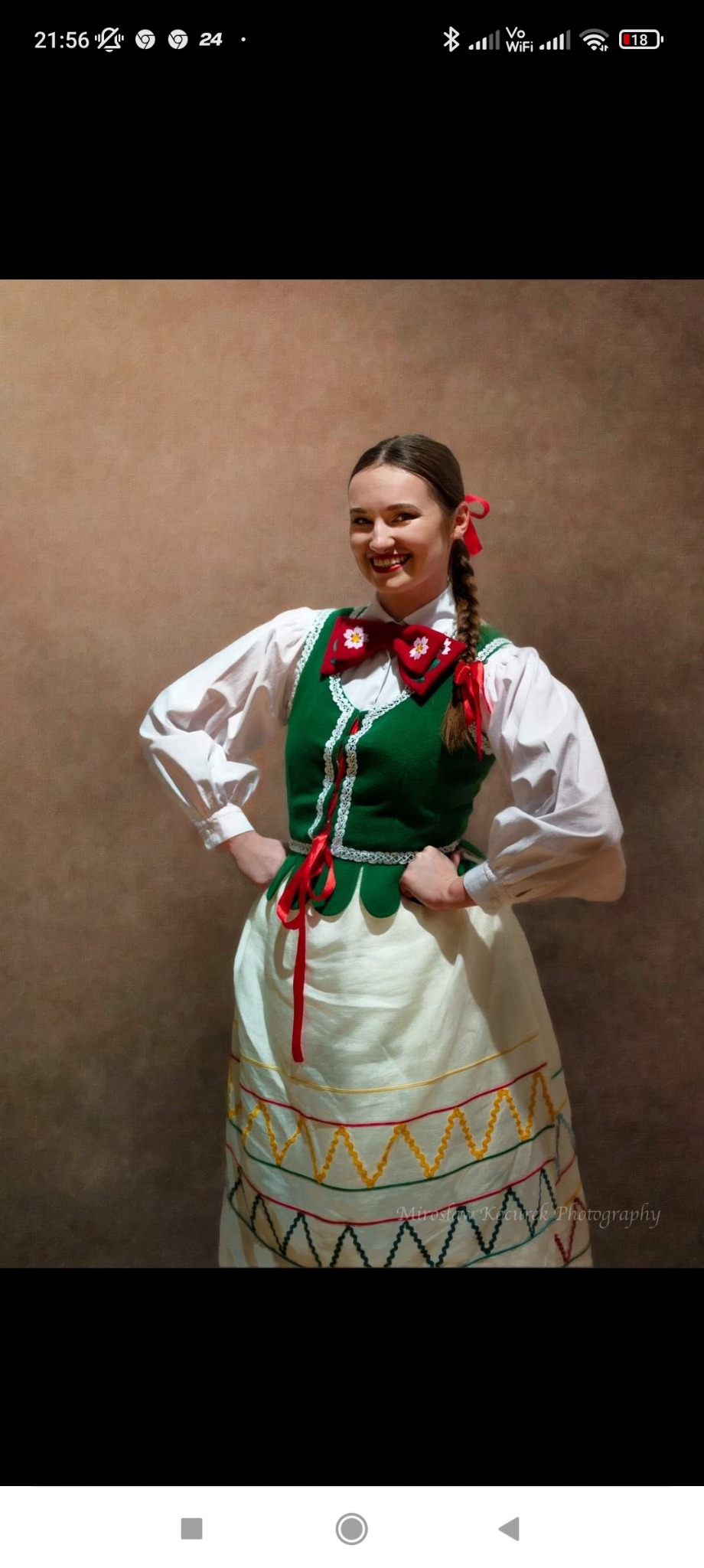 Uśmiechnięta młoda kobieta w ludowym stroju z zielonym gorsetem, białą bluzką i spódnicą z kolorowym haftem, pozująca na brązowym tle.