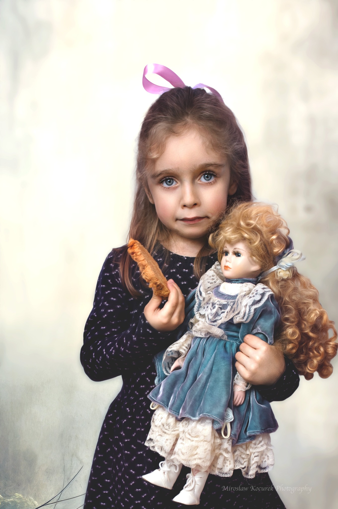 Dziewczynka z niebieskimi oczami trzyma w ramionach lalkę w aksamitnej sukience z koronką, w drugiej ręce trzyma ciastko.