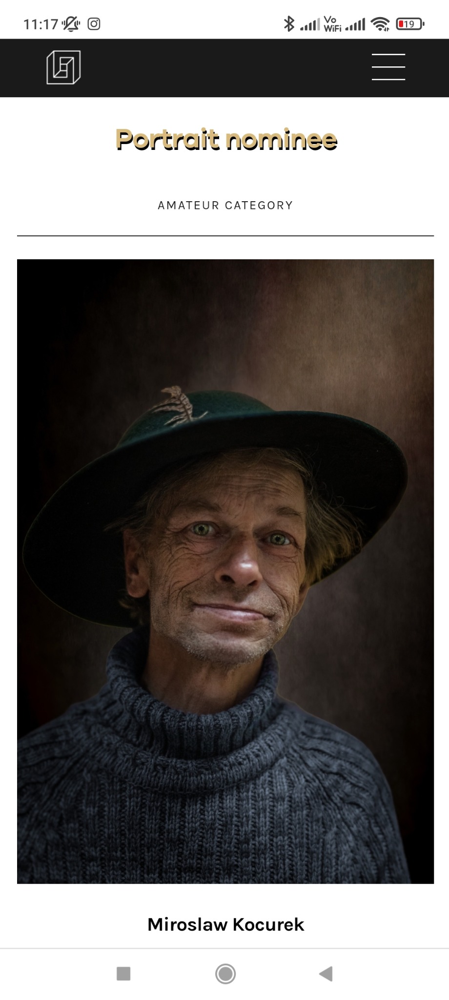 Portret starszego mężczyzny w ciemnym swetrze i kapeluszu ozdobionym piórem, uśmiechającego się lekko do kamery, na ciemnym tle.